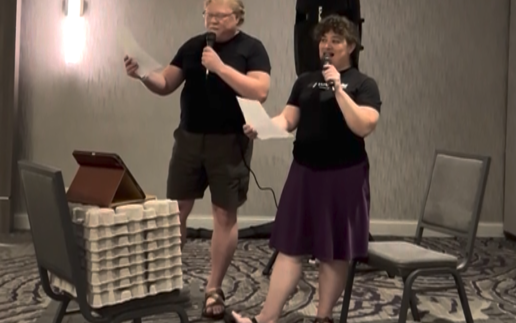 Jeff Gamet and Kelly Guimont with microphones in hand stand in front of an iPad and sing the song NosillaCast Island (modified version of Gilligan's Island)