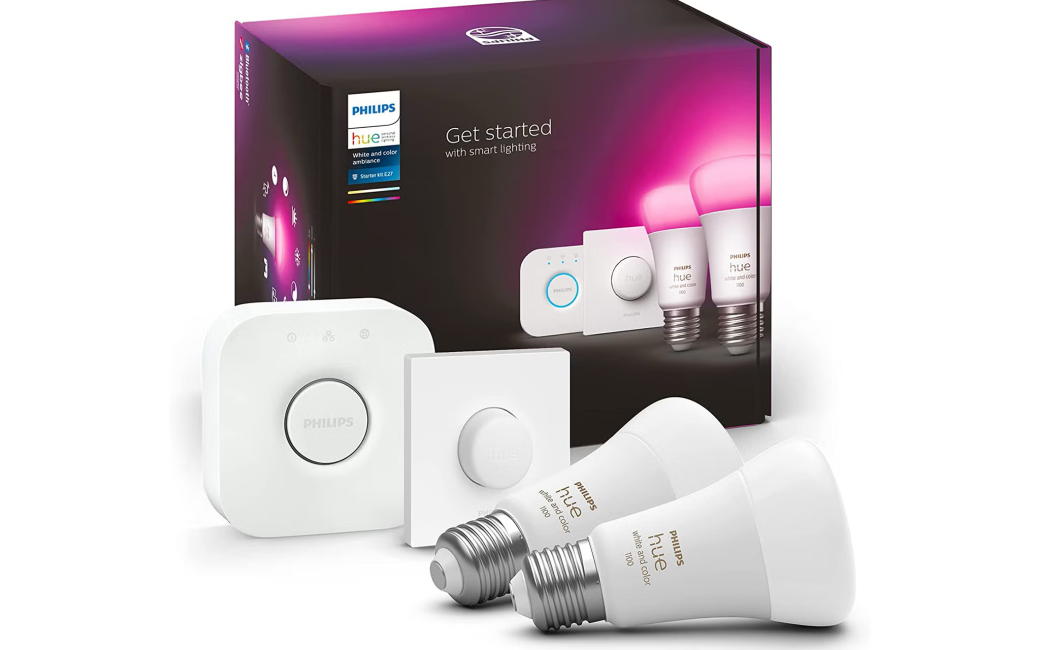 A product image of a Philips Hue Starter Kit. It shows a large black box with product imagery on the front. In front of the box are its contents. These include two bulbs, a dimmer knob, and a bridge.