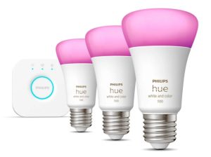 A product image of three Philips Hue bulbs and a Hue Bridge. The bulbs are lined up on an angle receding from the viewer and to the left, towards the bridge in the background. The bulbs are mostly white, lit up in a purple colour. The bridge is white with a lit up blue ring.