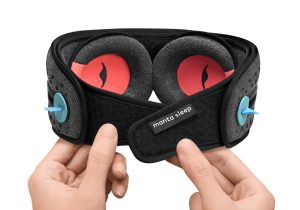 A picture of the Manta SOUND Sleep Mask from behind. It is shown held by a pair of hands. The hands are adjusting the velcro-like fastening at the back. Inside the mask you can see the C-shaped eye cups which have an orange-red interior.