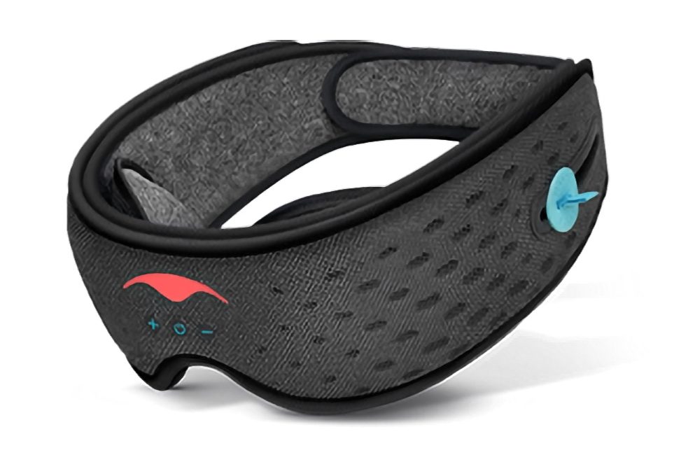 A product image showing the Manta SOUND Sleep Mask. It is a band of dark grey fabrics, wider in the front with an arch for the nose. There is a red Manta Sleep symbol — evocative of a Manta Ray — on the front, and a turquoise circle is visible on the near side which is the outer part of the Bluetooth speaker on that side. The mask is angled as if it is propped up at the back on a small box, however none is visible.