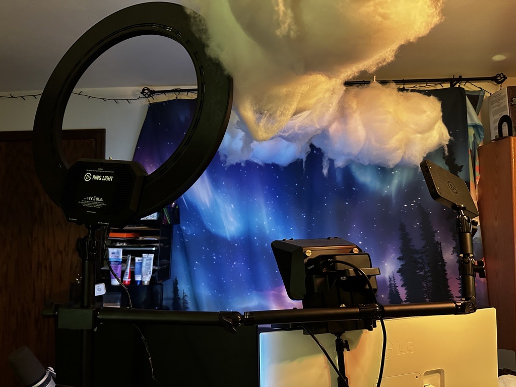 Back view of the two displays showing the ring light, teleprompter/camera combo and key light mini. You can also see her puffy clouds hanging from the ceiling that are sound dampeners, and a screen behind where she sits showign the northern lights and pine trees.