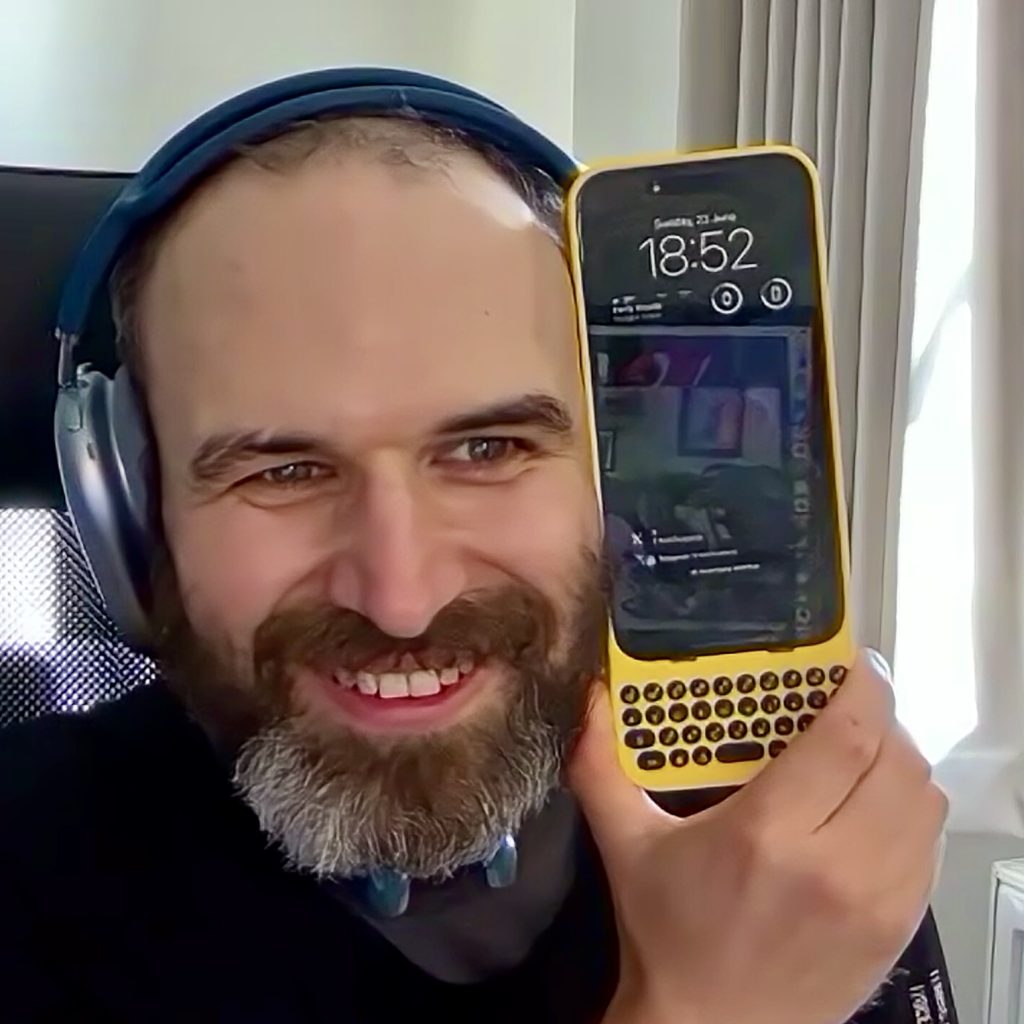 A bearded man wearing blue headphones holding an iPhone with a bright yellow keyboard case next to his head. The phone with case is as tall as his head.