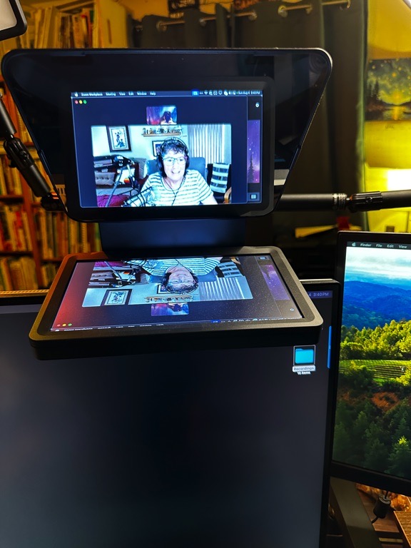 A photo of a teleprompter setup, displaying a video call on the screen. The visible setup includes the main monitor showing a person with headphones sitting in a home office environment. Below the teleprompter screen is another monitor displaying various computer applications and a scenic background wallpaper on a secondary screen.