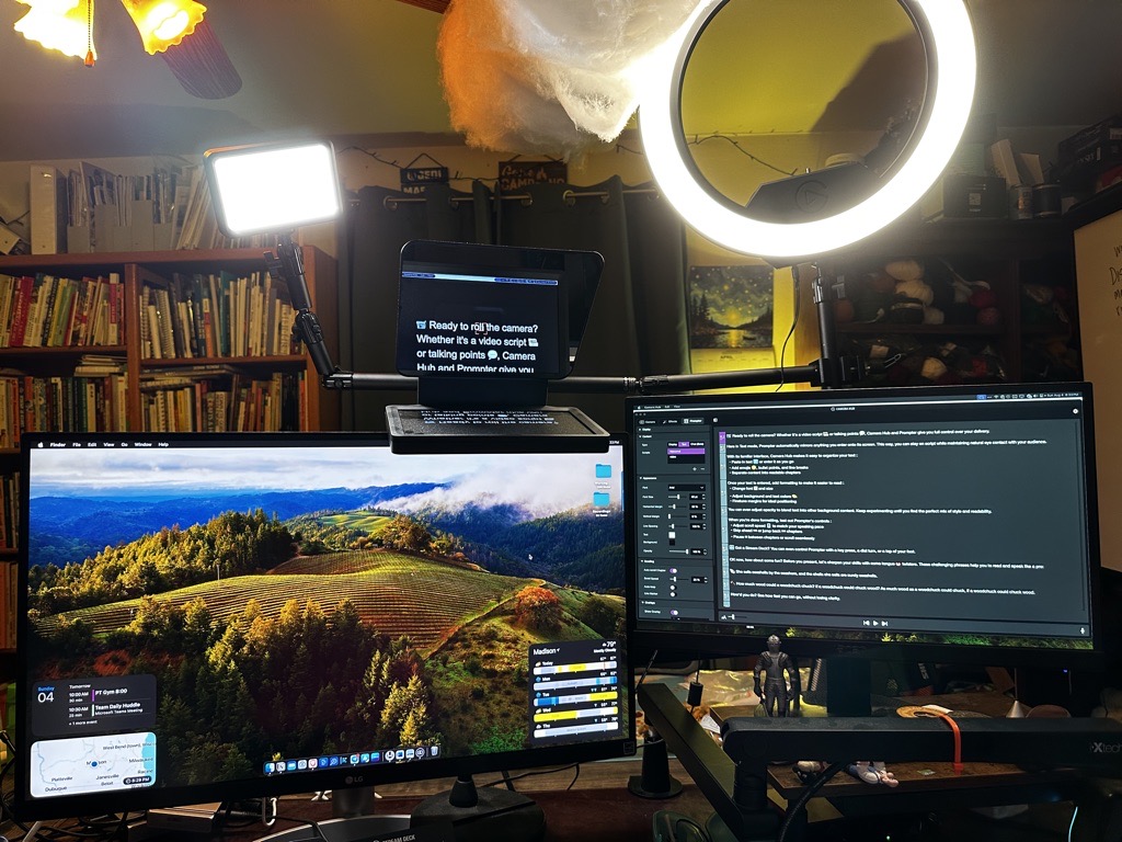 A comprehensive home office setup featuring multiple monitors and professional lighting equipment. The central focus is on a teleprompter displaying a script, flanked by a main monitor with a scenic wallpaper and another monitor displaying programming software. Additional accessories include a ring light for video conferencing and a small key light on a stand, enhancing the workspace for optimal video and photo capture.