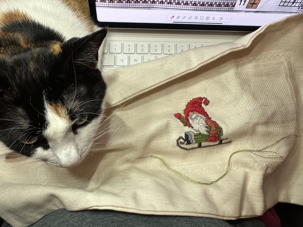 calico cat on my lap while cross stitching Christmas gnome with iPad in the background showing the pattern.