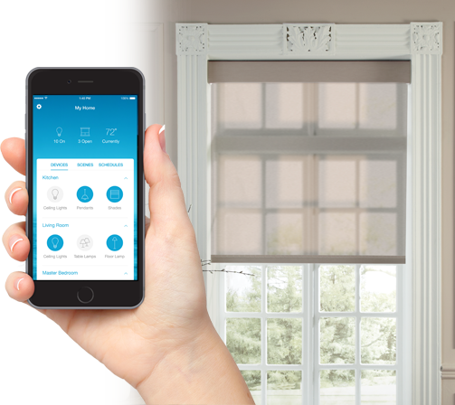 A hand holds a mobile phone with the face showing the Lutron mobile app with controls for devices, scenes, and schedules. Behind the phone is a window that has a light-filtering shade covering the upper half of the window. The shade is translucent and is light sand in color The window has an ornate white frame and white-rimmed window panes.